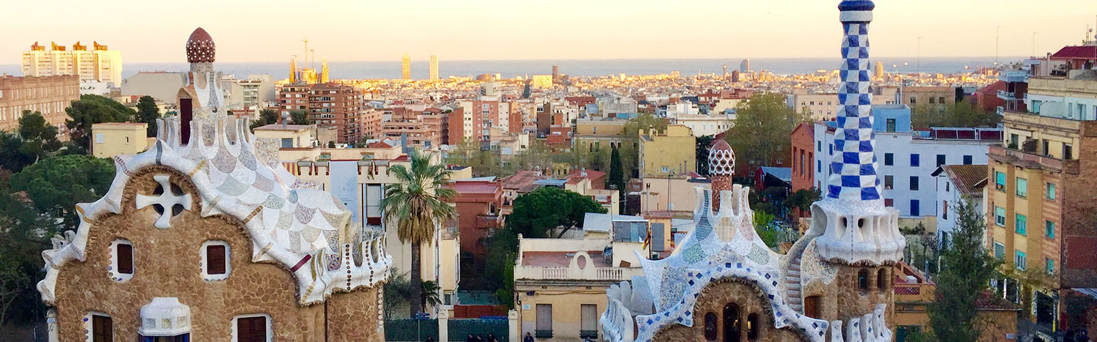 Two stone buildings with fancy, colorful roofs. An expansive view of Barcelona and the sea takes up the background.